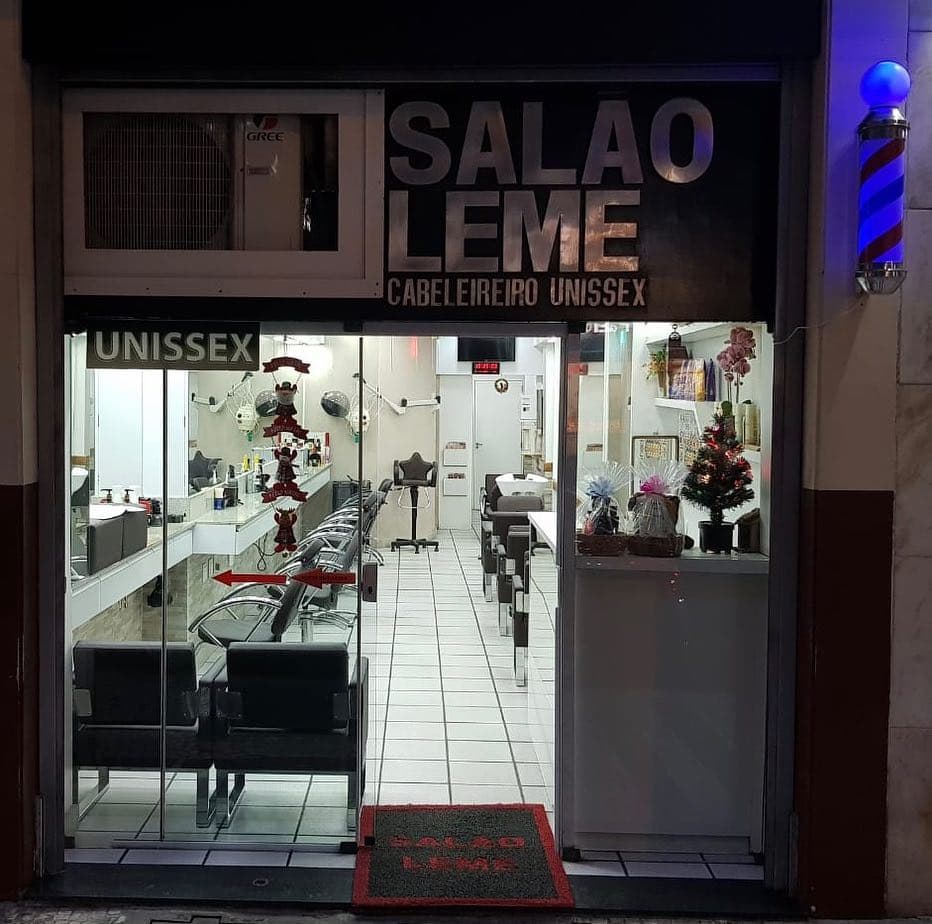 Fachada do Salão Leme à noite: letreiro preto com letras de aço, poste de barbeiro aceso, porta de vidro e capacho vermelho com o nome