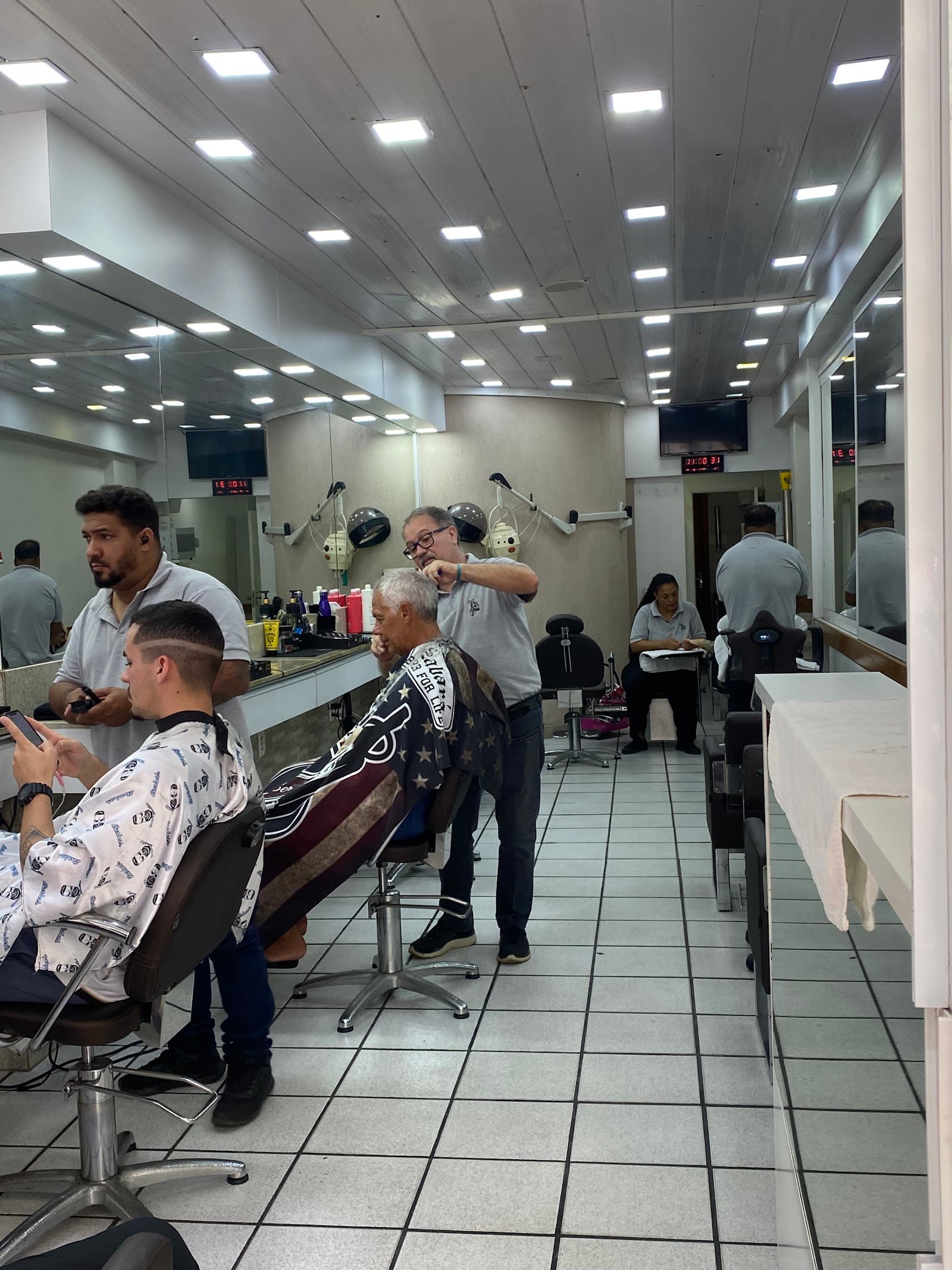 Cabeleireiro de óculos cortando o cabelo branco de um cliente, ao lado de um barbeiro fazendo degradê, no salão de piso de azulejo